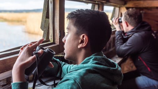 Young bird watcher and NT ranger sitting in the bird hide at Park End Moss wetland. They each have a pair of binoculars.